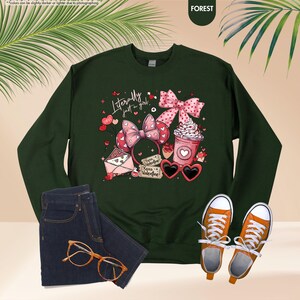 May include: Dark green sweatshirt with the text "Literally just a girl" and Valentine's Day graphics. The image includes a pair of blue jeans, orange trainers, and gold-rimmed glasses.