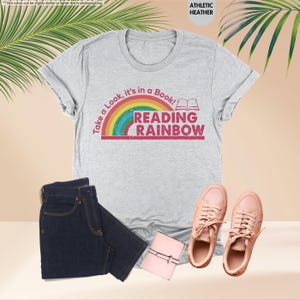 May include: A light gray athletic heather t-shirt with the text "Take a Look, it's in a Book! READING RAINBOW" in a rainbow design. The shirt is paired with dark blue jeans, pink sneakers, and a small pink wallet.