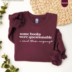 May include: A maroon long-sleeve crewneck sweatshirt featuring white text that reads some books were questionable i read them anyway with a small heart icon at the end. The garment has a ribbed collar, cuffs, and hem, and is made of fabric.
