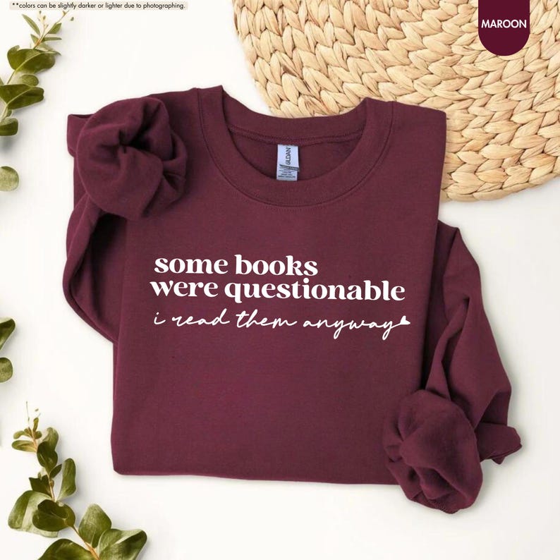 May include: A maroon long-sleeve crewneck sweatshirt featuring white text that reads some books were questionable i read them anyway with a small heart icon at the end. The garment has a ribbed collar, cuffs, and hem, and is made of fabric.