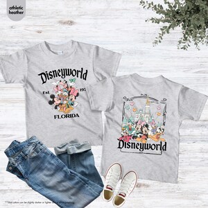 May include: Two gray t-shirts with Disneyworld graphics, jeans, and white sneakers. One shirt features Mickey, Minnie, and other characters with "Disneyworld Est 1971 Florida." The other shirt has a castle and characters with "Disneyworld 1971."