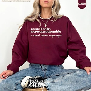May include: A maroon long-sleeve crewneck sweatshirt featuring white text that reads some books were questionable i read them anyway. The garment has a white inner collar trim and is paired with blue denim jeans and black and white sneakers.