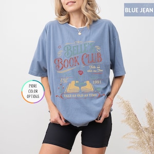 May include: A blue jean colored t-shirt with the text "Belle's Book Club" in a vintage design. The design includes the words "Tale as old as Time", "Est. 1991", and an open book illustration. The shirt has short sleeves.