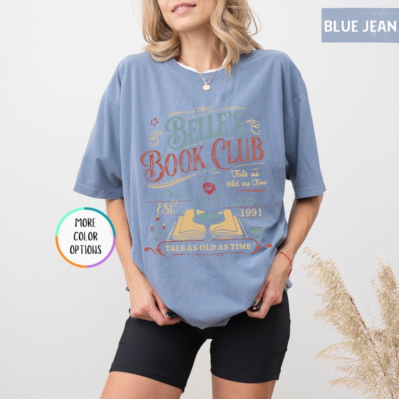 May include: A blue jean colored t-shirt with the text "Belle's Book Club" in a vintage design. The design includes the words "Tale as old as Time", "Est. 1991", and an open book illustration. The shirt has short sleeves.