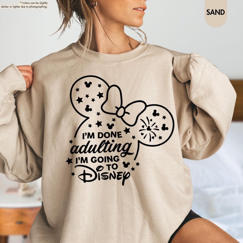 May include: A tan sweatshirt with a black graphic of a Mickey Mouse head with a bow and fireworks. The text "I'm done adulting I'm going to Disney" is printed below the graphic.