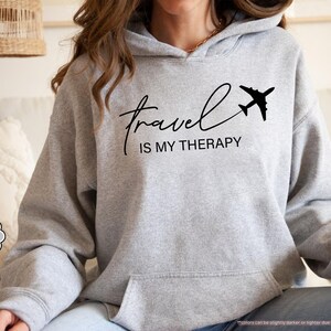 Pu&ograve; includere: Felpa con cappuccio grigio chiaro con la scritta "Travel is my Therapy" in corsivo nero e una grafica di aereo nero. La felpa con cappuccio ha un cappuccio con coulisse e una tasca frontale. Le parole "Sport Grey" sono in un piccolo cerchio.