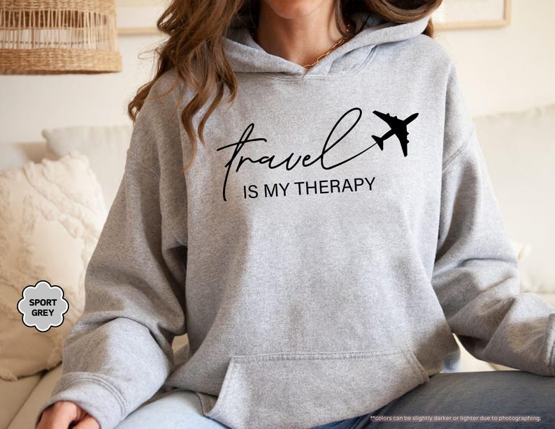 Pu&ograve; includere: Felpa con cappuccio grigio chiaro con la scritta "Travel is my Therapy" in corsivo nero e una grafica di aereo nero. La felpa con cappuccio ha un cappuccio con coulisse e una tasca frontale. Le parole "Sport Grey" sono in un piccolo cerchio.