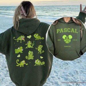 Op de afbeelding: Een olijfgroene hoodie met een cartoonontwerp van Pascal de kameleon uit de film Rapunzel. De achterkant van de hoodie heeft meerdere Pascal-illustraties. De voorkant van de hoodie heeft de naam "PASCAL" en het jaar "2010".