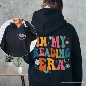 a woman wearing a black hoodie with the words in my reading era printed on
