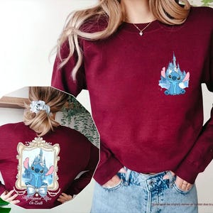 May include: Maroon sweatshirt featuring a blue cartoon character and castle design. The front has a small graphic, while the back displays a larger framed image with the text "The Happiest Place On Earth".