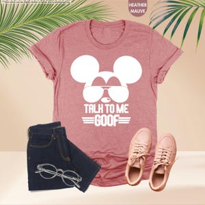 May include: Heather mauve t-shirt with a white Mickey Mouse graphic wearing sunglasses and the text "TALK TO ME GOOF". The image also includes dark blue jeans, eyeglasses, and pink trainers.