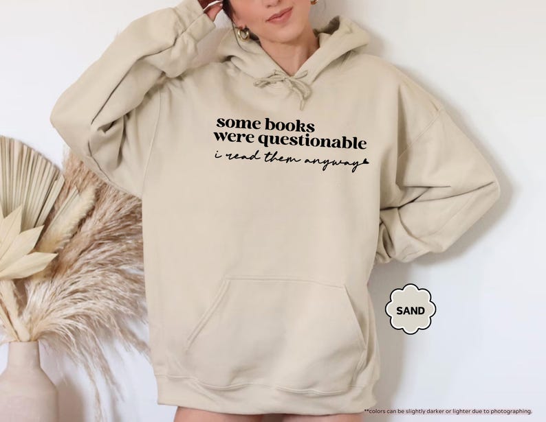 May include: A sand-colored hooded sweatshirt featuring a front kangaroo pocket and a drawstring hood. The chest displays black text that reads: some books were questionable i read them anyway, with a small heart icon at the end of the sentence.