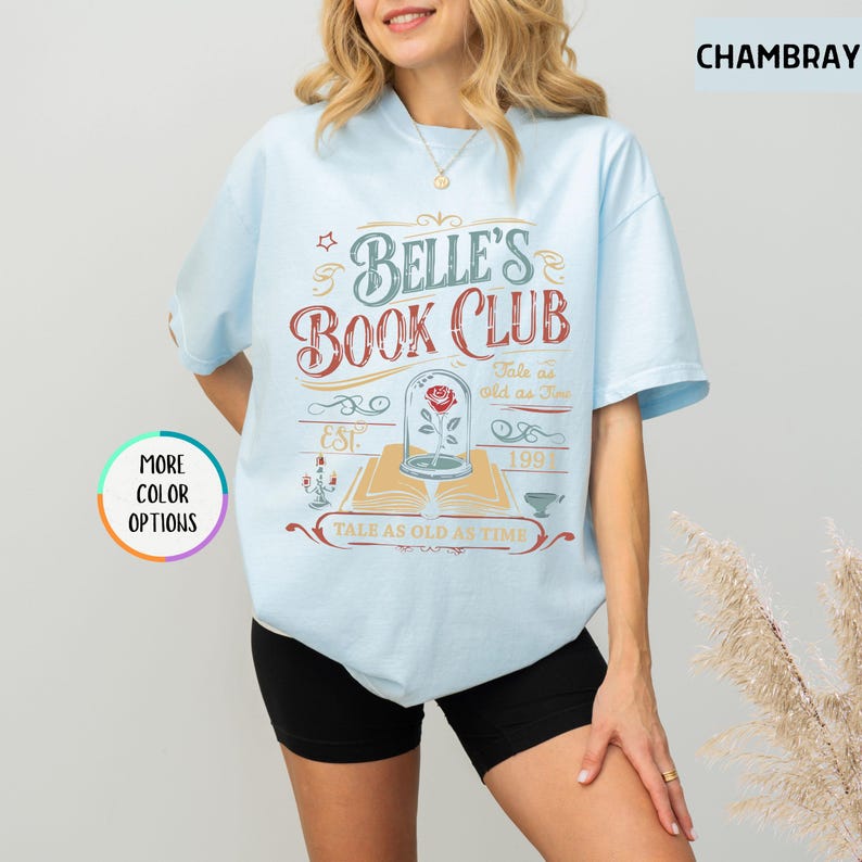 May include: Light blue t-shirt with the text "BELLE'S BOOK CLUB" in a vintage design. The graphic includes a rose in a glass dome, an open book, and the phrase "Tale as Old as Time". The shirt also has the text "EST. 199" and "MORE COLOR OPTIONS".