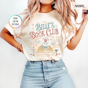 May include: Ivory-colored t-shirt with the text "Belle's Book Club" in a vintage design. The graphic includes a rose in a glass dome, an open book, and the phrase "Tale as old as time."