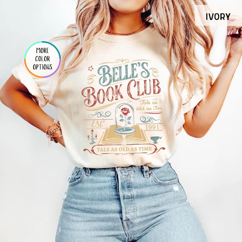 May include: Ivory-colored t-shirt with the text "Belle's Book Club" in a vintage design. The graphic includes a rose in a glass dome, an open book, and the phrase "Tale as old as time."