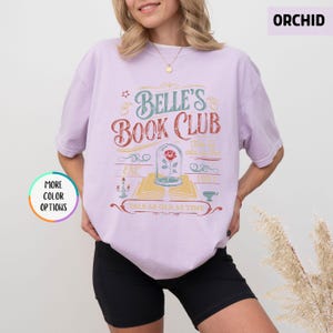 May include: Orchid-colored t-shirt with the text "Belle's Book Club" in a vintage design. The graphic includes a rose in a glass dome, an open book, and the phrase "Tale as Old as Time". The shirt has short sleeves.