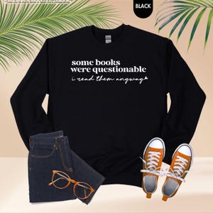 May include: A black long-sleeve crewneck sweatshirt featuring the white text 'some books were questionable i read them anyway' with a small heart icon at the end. The garment is displayed alongside dark blue denim jeans, orange sneakers, and glasses.