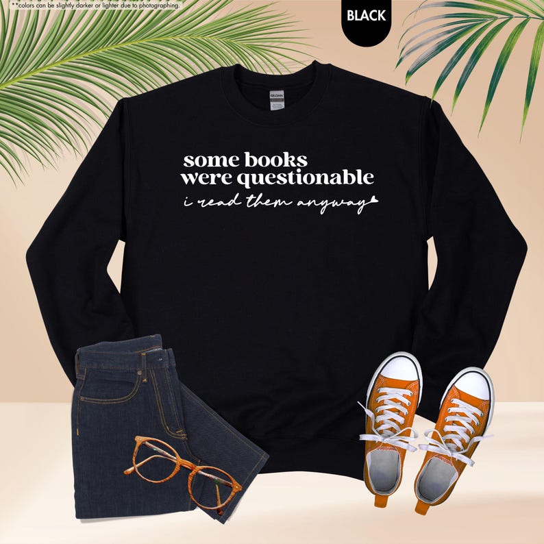 May include: A black long-sleeve crewneck sweatshirt featuring the white text 'some books were questionable i read them anyway' with a small heart icon at the end. The garment is displayed alongside dark blue denim jeans, orange sneakers, and glasses.