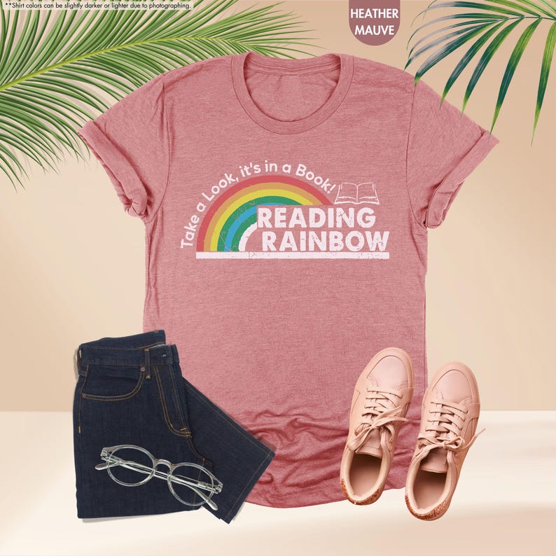 May include: Heather mauve t-shirt with the text "Take a Look, it's in a Book! READING RAINBOW" and a rainbow graphic. The image also shows a pair of dark blue jeans, clear glasses, and pink sneakers.