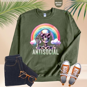 May include: Military green sweatshirt featuring a skeleton wearing heart-shaped sunglasses and a rainbow. The word "ANTISOCIAL" is printed below. The image also includes dark blue jeans, orange trainers, and eyeglasses.