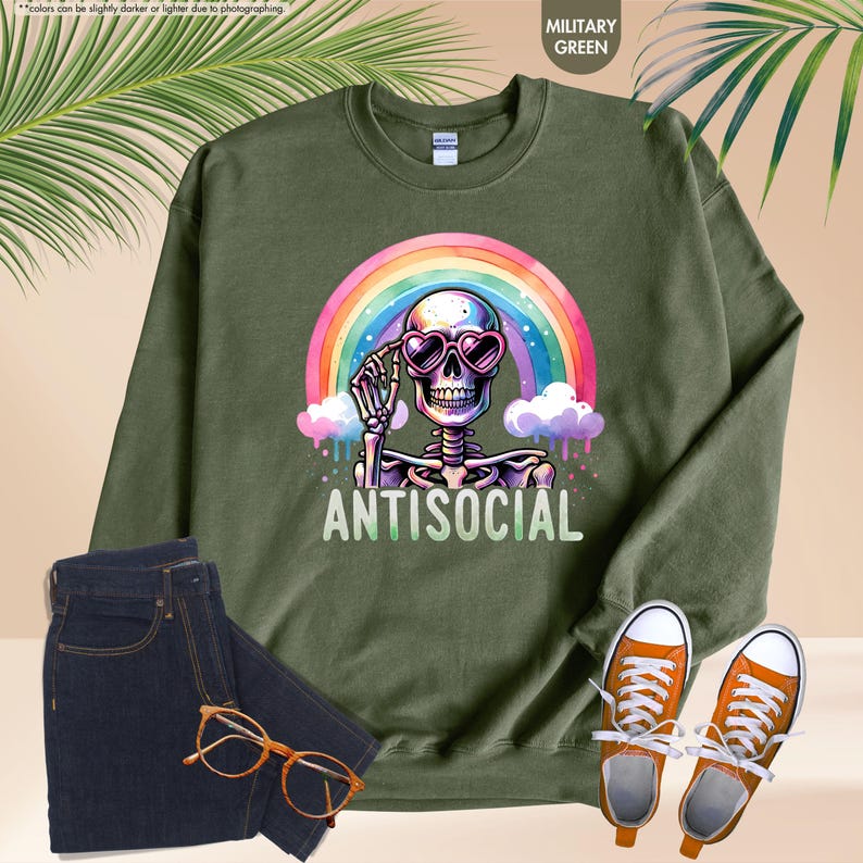 May include: Military green sweatshirt featuring a skeleton wearing heart-shaped sunglasses and a rainbow. The word "ANTISOCIAL" is printed below. The image also includes dark blue jeans, orange trainers, and eyeglasses.