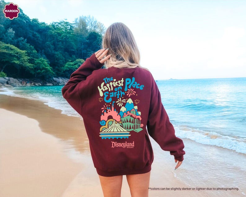 Pu&ograve; includere: Una felpa color granata con una stampa grafica di un castello dei cartoni animati e fuochi d'artificio. Il testo "The Happiest Place on Earth" e "Disneyland" sono stampati sulla felpa.