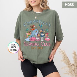 May include: A green t-shirt with a graphic of mice sewing on a Cinderella slipper. The text reads "Cinderella's Sewing Club Est. 1950".