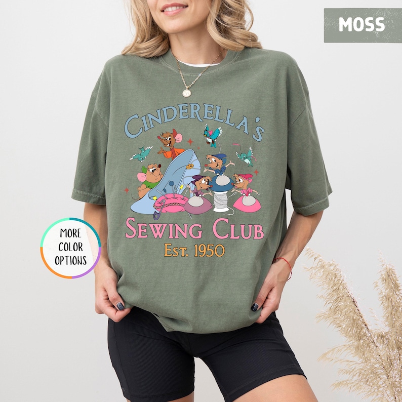 May include: A green t-shirt with a graphic of mice sewing on a Cinderella slipper. The text reads "Cinderella's Sewing Club Est. 1950".