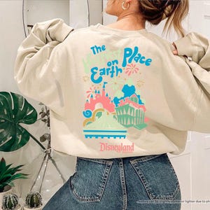 Pu&ograve; includere: Una felpa beige con una stampa grafica di un castello e fuochi d'artificio. Il testo "The Happiest Place on Earth" e "Disneyland" sono stampati sulla felpa.