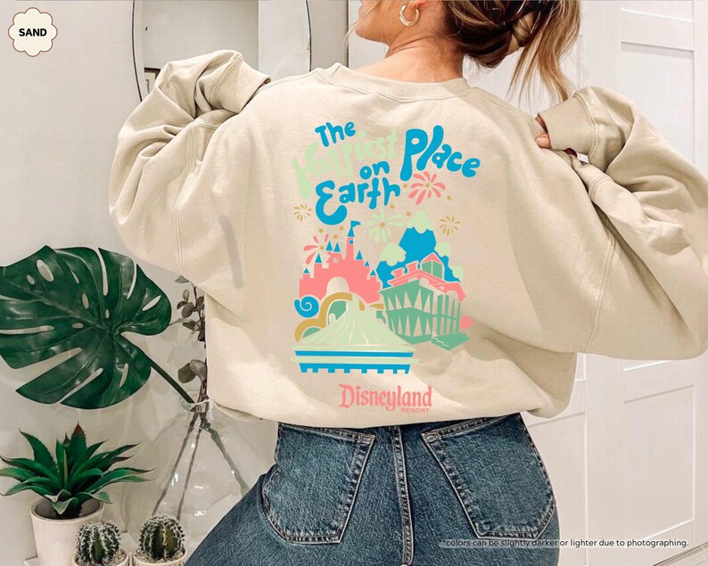 Pu&ograve; includere: Una felpa beige con una stampa grafica di un castello e fuochi d'artificio. Il testo "The Happiest Place on Earth" e "Disneyland" sono stampati sulla felpa.