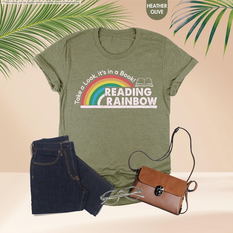 May include: Heather olive green t-shirt with the text "Take a Look, it's in a Book! READING RAINBOW" and a rainbow graphic. The shirt is paired with dark blue jeans, a brown purse, and eyeglasses. The shirt is a casual style.