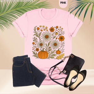 May include: Pale pink t-shirt featuring a floral and pumpkin design. The outfit includes dark wash jeans, a black handbag, and black flats. The word "PINK" is visible in the top right corner.