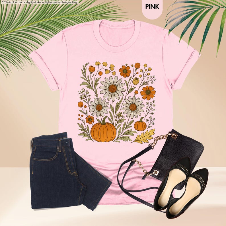 May include: Pale pink t-shirt featuring a floral and pumpkin design. The outfit includes dark wash jeans, a black handbag, and black flats. The word "PINK" is visible in the top right corner.