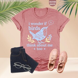 May include: A heather mauve t-shirt featuring a light blue graphic of a bird and the text "i wonder if birds think about me too" with a small thought bubble containing a heart, paired with dark blue denim jeans and light pink sneakers.