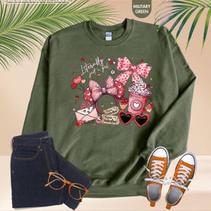 May include: Military green sweatshirt featuring the phrase "Literally just a girl" and Valentine's Day themed graphics. The design includes a pink bow, heart-shaped sunglasses, and a coffee cup. Shown with blue jeans, orange trainers, and glasses.