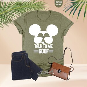 May include: Heather olive green t-shirt with a white Mickey Mouse graphic wearing sunglasses and the text "TALK TO ME GOOF". The image also includes a pair of dark blue jeans, a brown purse, and eyeglasses.