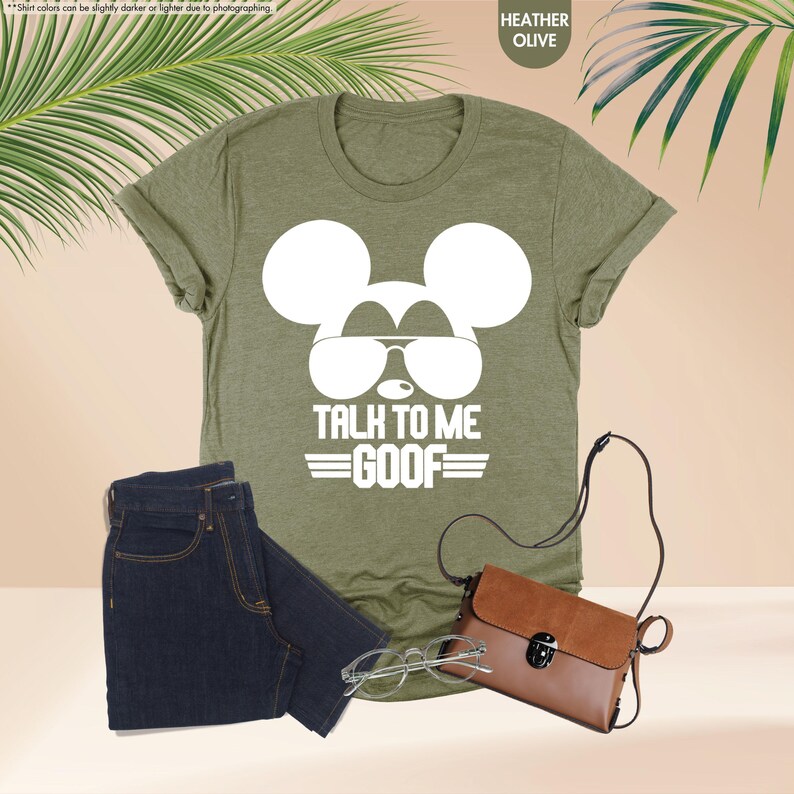May include: Heather olive green t-shirt with a white Mickey Mouse graphic wearing sunglasses and the text "TALK TO ME GOOF". The image also includes a pair of dark blue jeans, a brown purse, and eyeglasses.