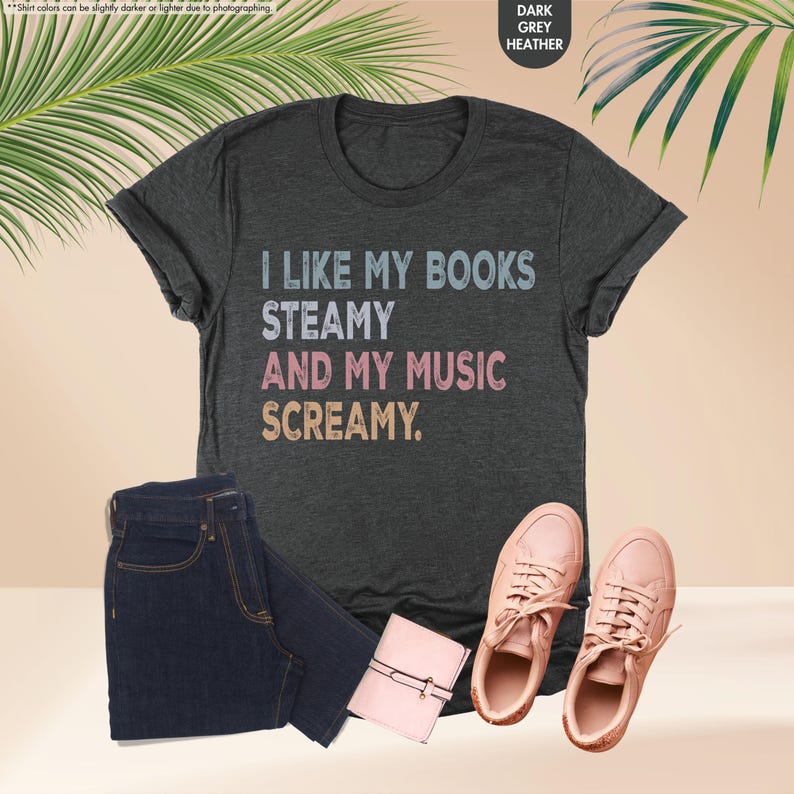 May include: Dark grey heather t-shirt with the text "I LIKE MY BOOKS STEAMY AND MY MUSIC SCREAMY." The image also includes a pair of dark wash jeans, a small pink wallet, and a pair of pink sneakers.