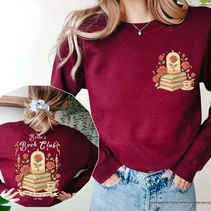 Puede incluir: Sudadera granate con un dise&ntilde;o floral que incluye una rosa, libros y una taza de t&eacute;. La parte delantera tiene un peque&ntilde;o gr&aacute;fico, mientras que la parte trasera muestra "Belle's Book Club" con el texto "Tale as Old as Time".