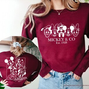 May include: A maroon sweatshirt featuring a white graphic of Mickey Mouse and friends, with the text "MICKEY & CO Est. 1928". The back of the sweatshirt displays a similar design with Mickey Mouse characters. The sweatshirt is made of a soft material.
