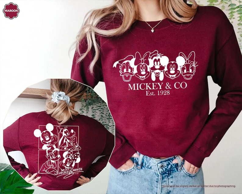May include: A maroon sweatshirt featuring a white graphic of Mickey Mouse and friends, with the text "MICKEY & CO Est. 1928". The back of the sweatshirt displays a similar design with Mickey Mouse characters. The sweatshirt is made of a soft material.
