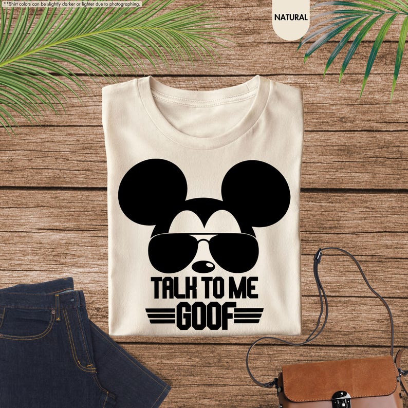 May include: Cream-coloured t-shirt with a black graphic of Mickey Mouse wearing sunglasses. The text "TALK TO ME GOOF" is below the image, with horizontal lines above and below. The shirt is displayed on a wooden surface.