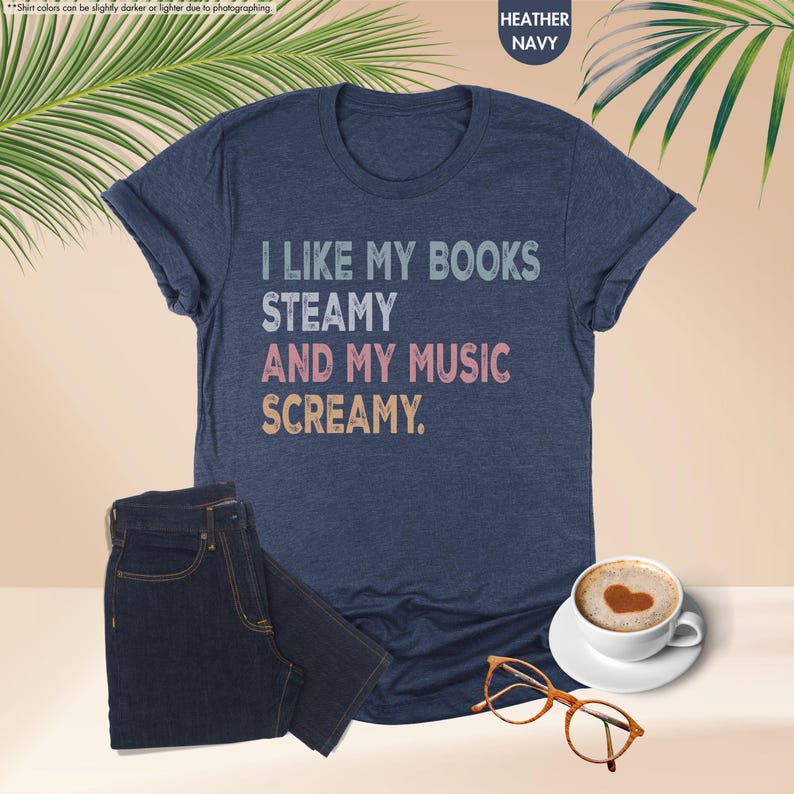 May include: Heather navy t-shirt with the text "I LIKE MY BOOKS STEAMY AND MY MUSIC SCREAMY." The shirt is paired with dark wash jeans, a cup of coffee with a heart design, and brown eyeglasses. The shirt is a casual wear item.