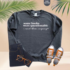May include: A dark heather gray crewneck sweatshirt featuring the white text some books were questionable i read them anyway, paired with dark blue denim jeans, orange canvas sneakers with white laces, and brown tortoise shell eyeglasses.