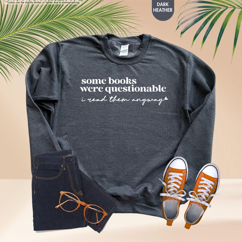 May include: A dark heather gray crewneck sweatshirt featuring the white text some books were questionable i read them anyway, paired with dark blue denim jeans, orange canvas sneakers with white laces, and brown tortoise shell eyeglasses.