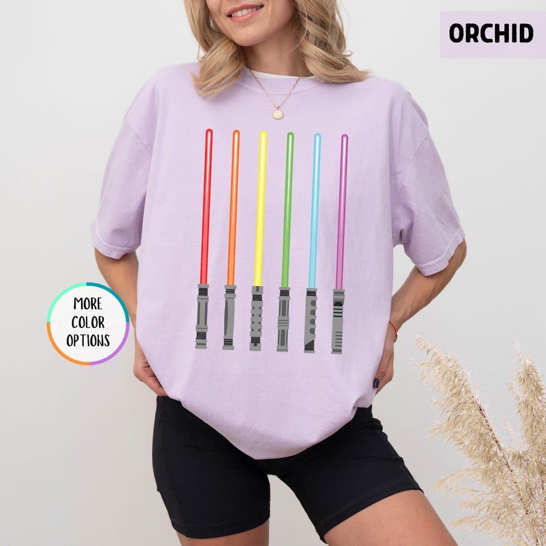 K&ouml;nnte beinhalten: Ein helllila T-Shirt mit sechs farbigen Lichtschwertern in Rot, Orange, Gelb, Gr&uuml;n, Blau und Lila. Das Wort "ORCHID" ist in einem lila Rechteck aufgedruckt. Das Shirt hat auch einen "MORE COLOR OPTIONS"-Button.