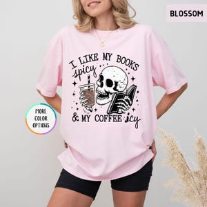 May include: Pale pink t-shirt featuring the phrase "I like my books spicy & my coffee icy" alongside a skull graphic holding a book and a coffee. The tee has a loose fit and short sleeves.