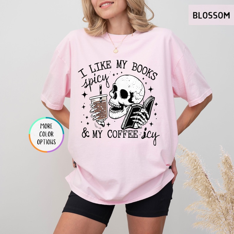 May include: Pale pink t-shirt featuring the phrase "I like my books spicy & my coffee icy" alongside a skull graphic holding a book and a coffee. The tee has a loose fit and short sleeves.