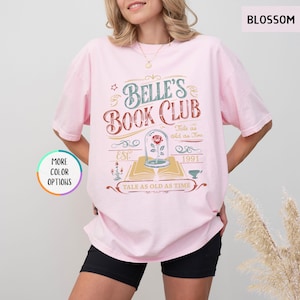 May include: A light pink t-shirt with the text "Belle's Book Club" in a vintage design. The graphic includes a rose in a glass dome, the words "Tale as old as Time", and the year "1991". The shirt also has the word "Blossom" printed on it.