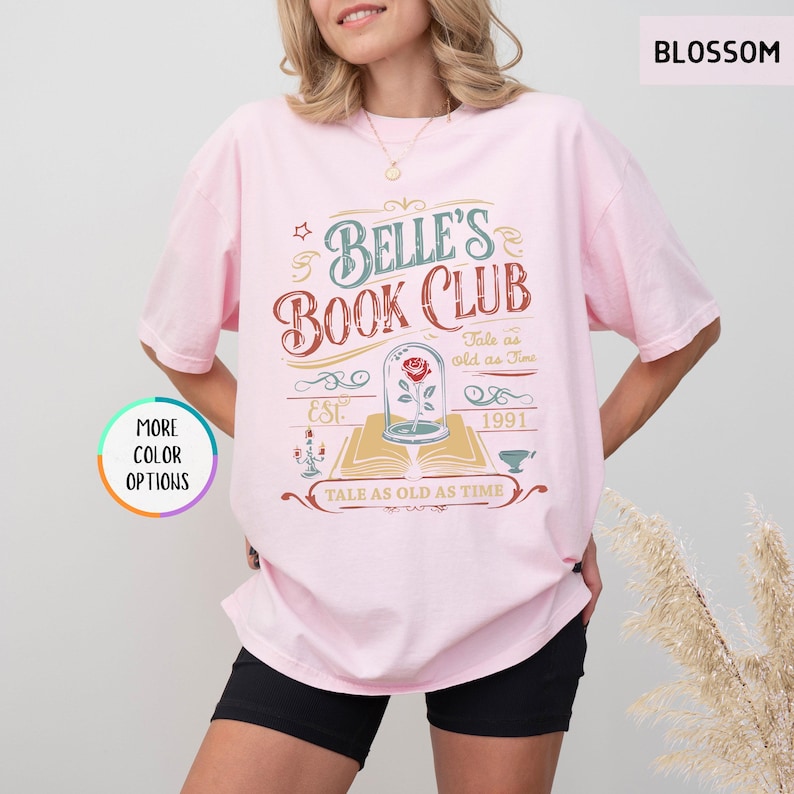 May include: A light pink t-shirt with the text "Belle's Book Club" in a vintage design. The graphic includes a rose in a glass dome, the words "Tale as old as Time", and the year "1991". The shirt also has the word "Blossom" printed on it.